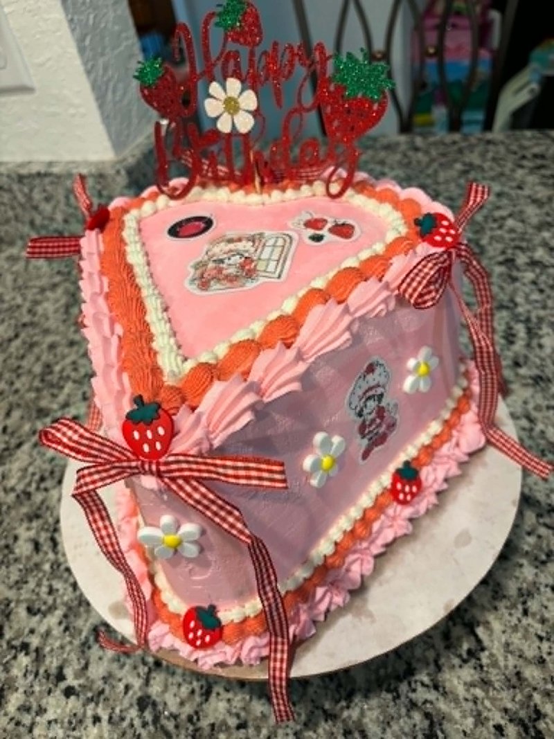 Heart-shaped pink buttercream cake with Strawberry Shortcake characters, red gingham bows, strawberry accents, and a Happy Birthday topper
