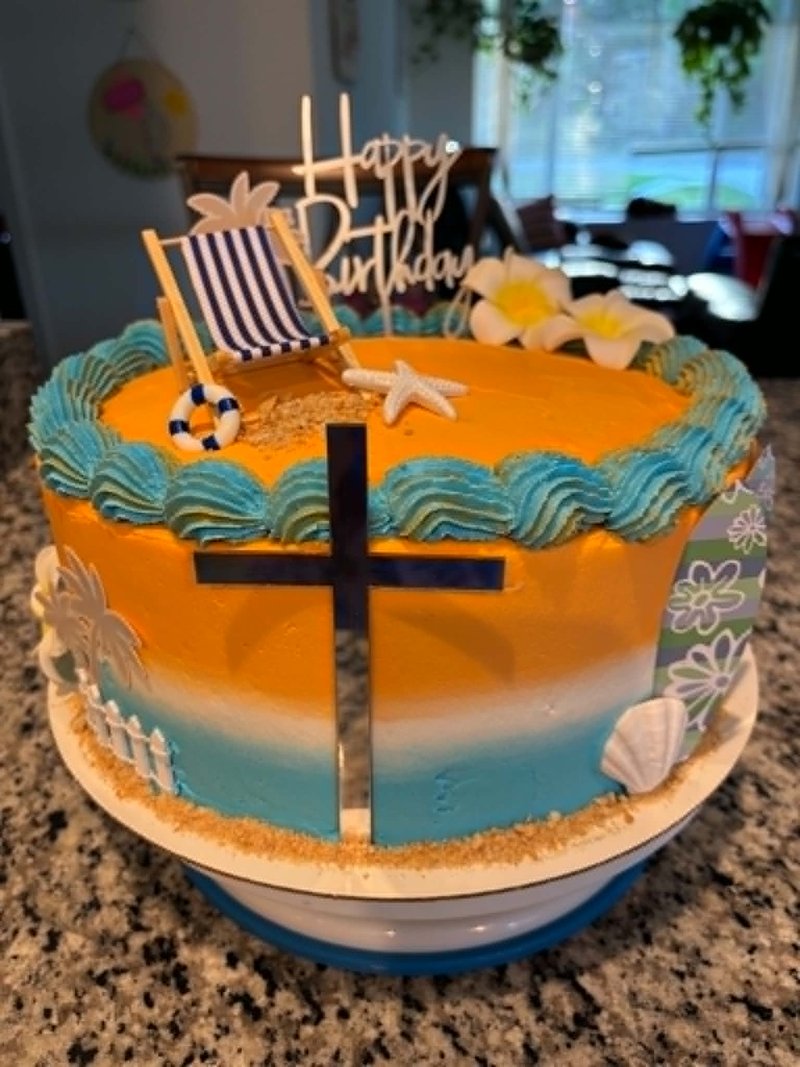 Orange-and-teal sunset gradient cake with a beach-chair topper, starfish, plumeria flowers, sandy base, and a cross with Happy Birthday lettering