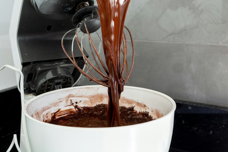 Stand mixer with whisk attachment pouring chocolate batter into a white bowl on a marble counter
